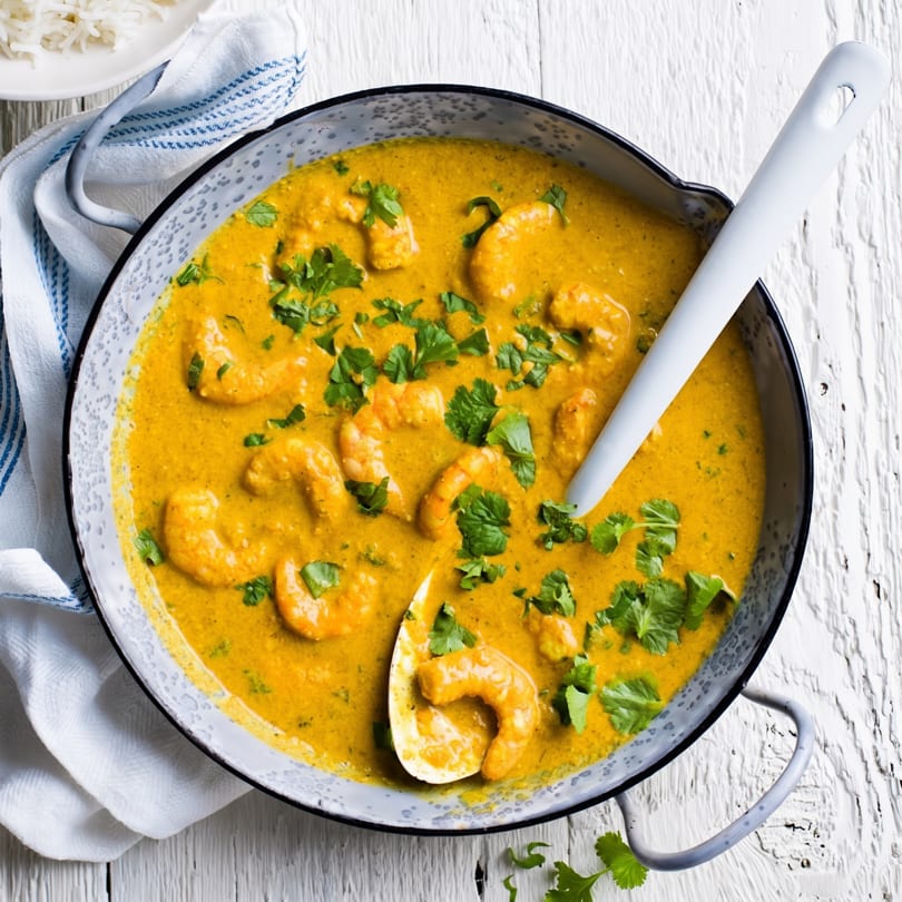 Shrimp curry in a gray pot with a white spoon, garnished with cilantro, next to white rice.
