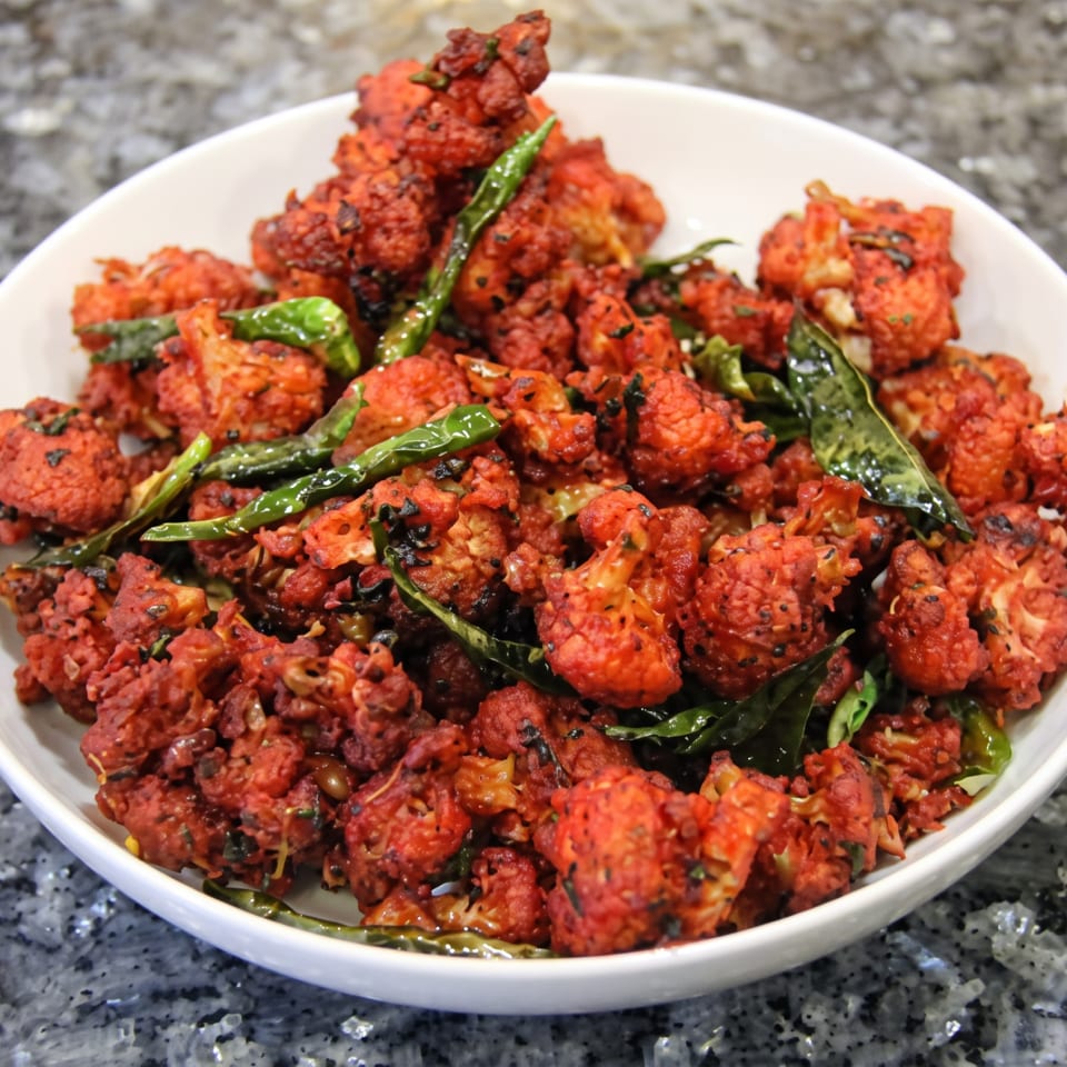 A white bowl filled with vibrant red fried cauliflower florets, garnished with green chilies and curry leaves.