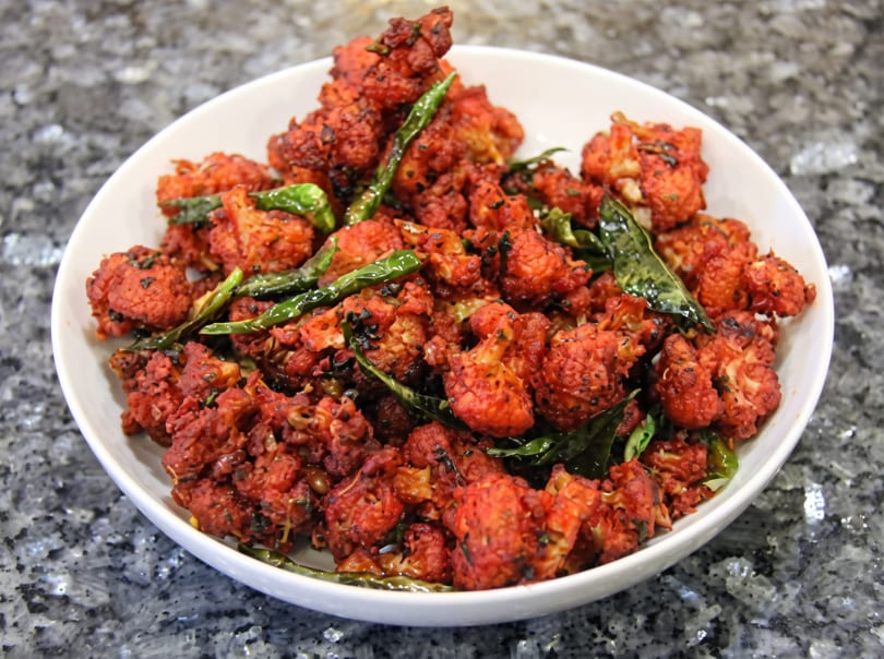 A white bowl filled with vibrant red fried cauliflower florets, garnished with green chilies and curry leaves.
