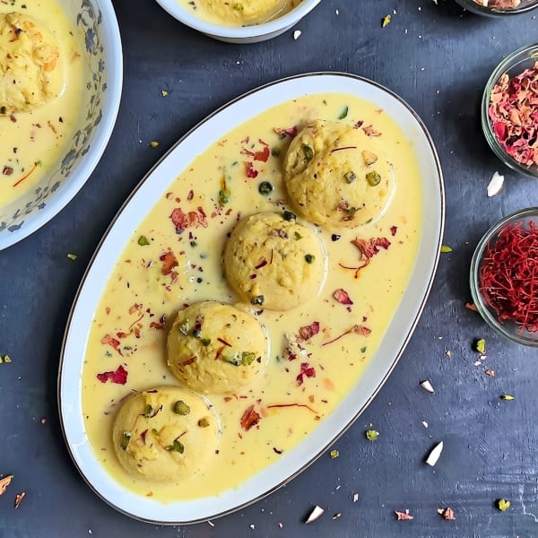 Rasmalai, a dessert with four yellow dumplings in creamy sauce, garnished with rose petals and nuts.