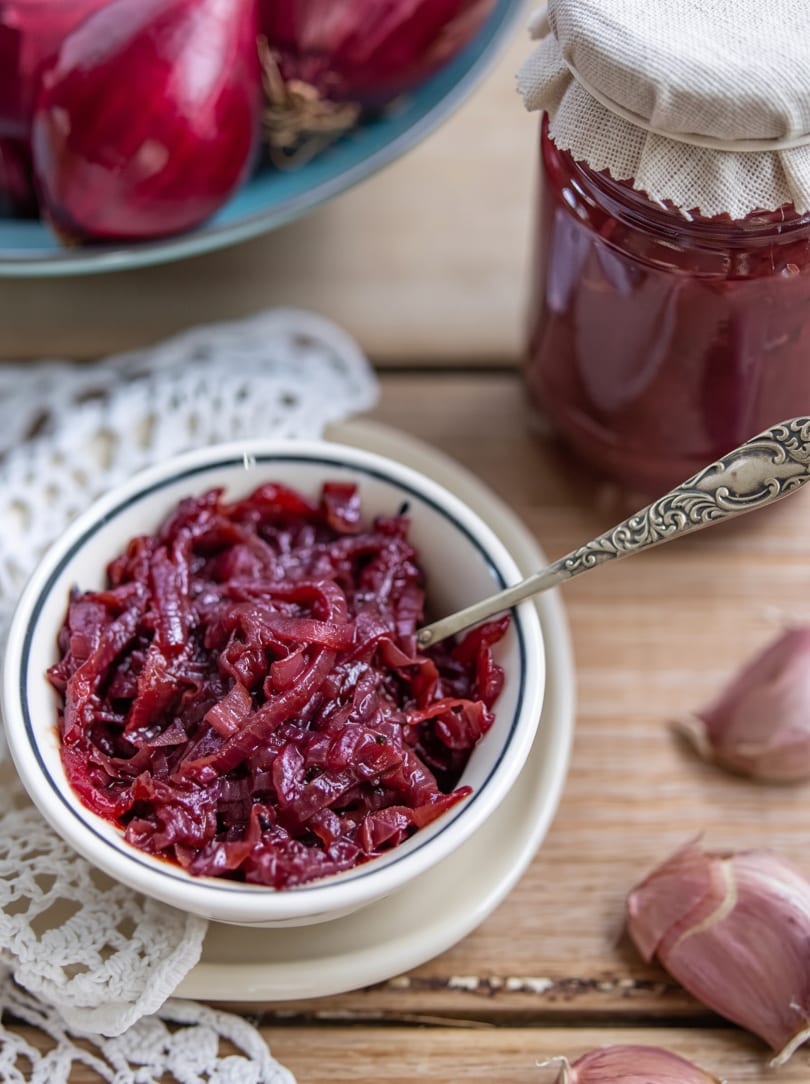 A bowl of red onion jam with a spoon, a jar, and fresh onions on a wooden table.