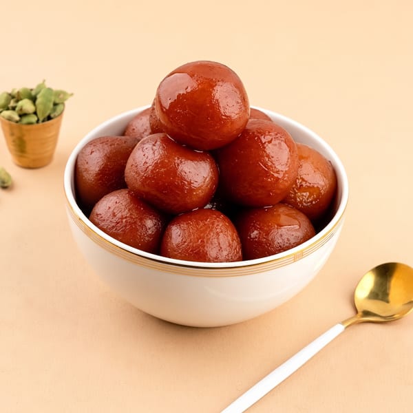 A white bowl filled with glistening brown gulab jamun, with a gold spoon and cardamom pods nearby.