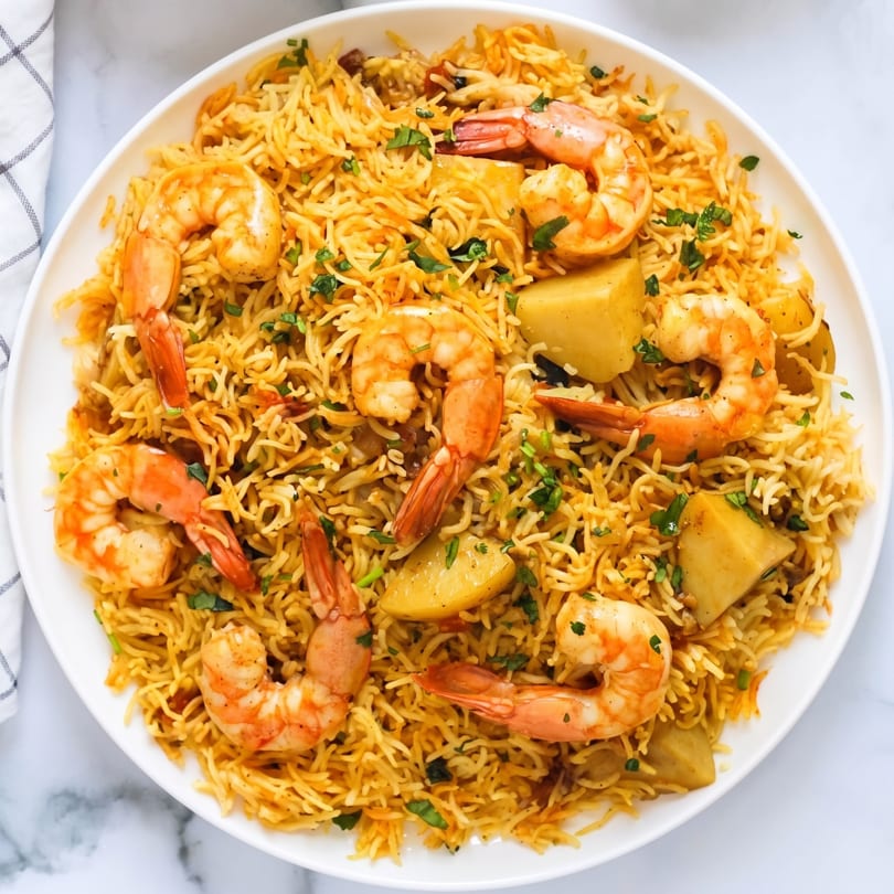 A white plate filled with shrimp biryani, featuring large shrimp, rice, potatoes, and fresh herbs.