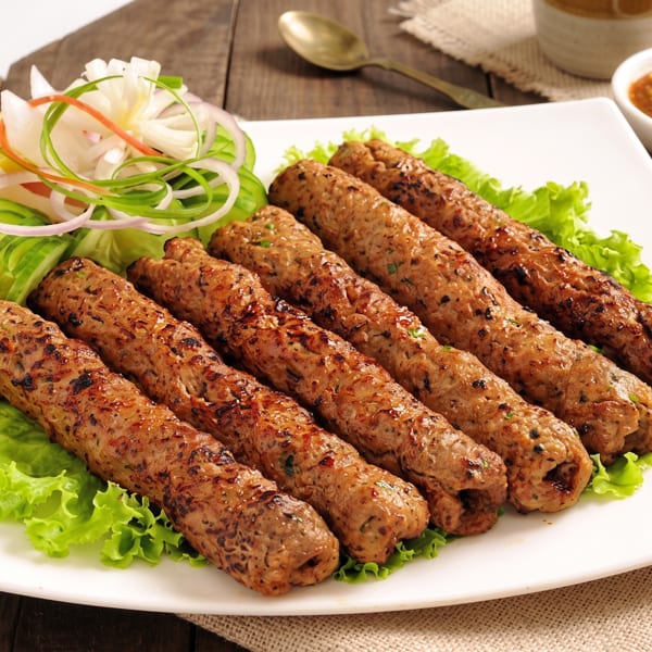 Six grilled seekh kebabs on a bed of lettuce with a side of salad and dipping sauce.