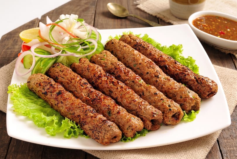 Six grilled seekh kebabs on a bed of lettuce with a side of salad and dipping sauce.