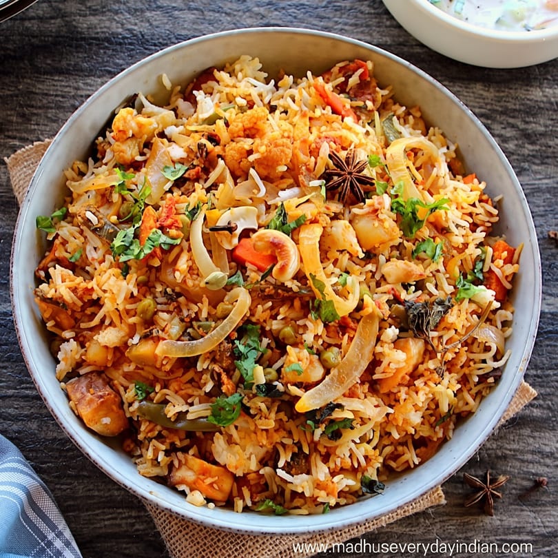 Close-up of a bowl of vegetable biryani with rice, vegetables, and spices on a wooden table.