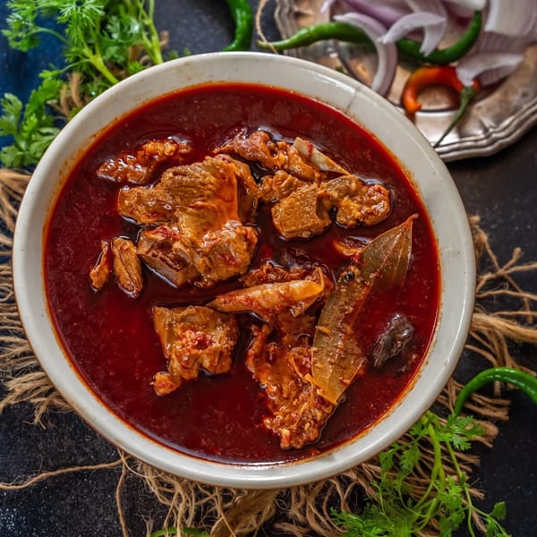 A bowl of rich red mutton curry with bay leaves, served with cilantro, onions, and chilies.