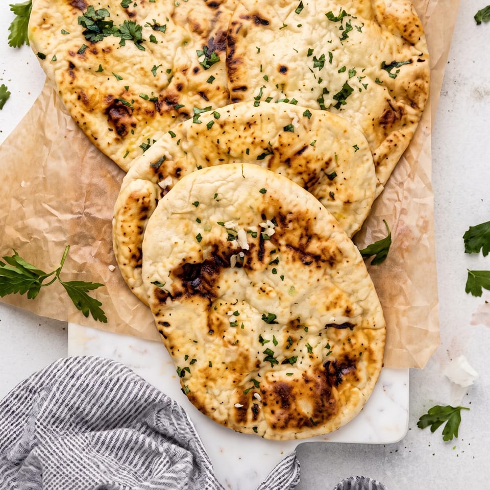 Four pieces of golden-brown naan bread with herbs on parchment paper, with a small bowl of dip.