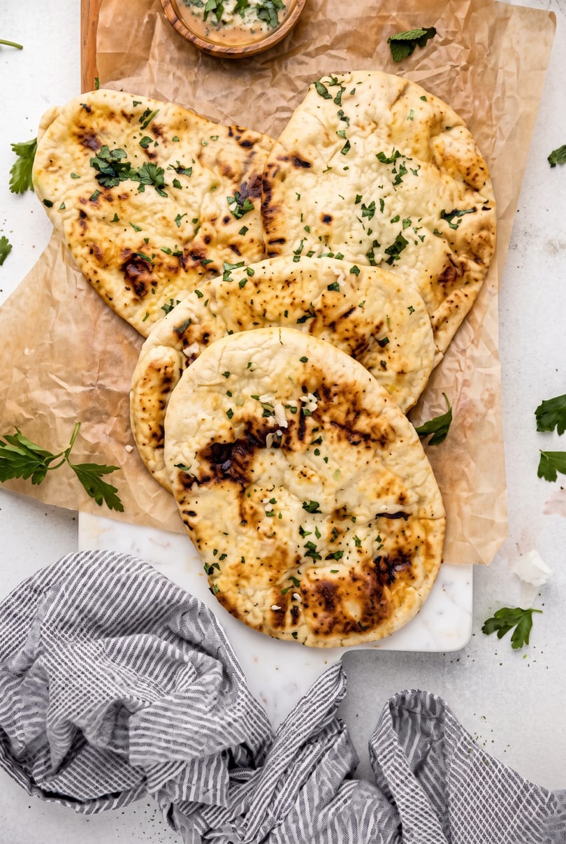 Four pieces of golden-brown naan bread with herbs on parchment paper, with a small bowl of dip.