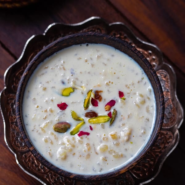 Creamy rice pudding dessert, garnished with pistachios, raisins, and rose petals, in a dark wooden bowl.