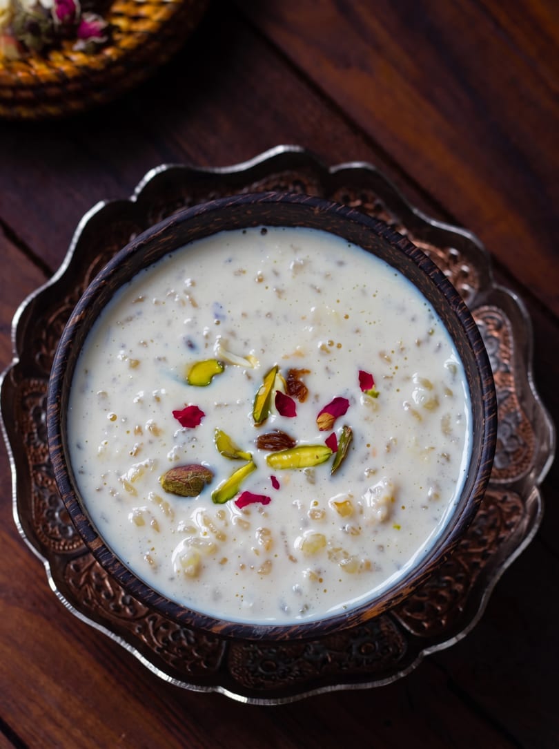 Creamy rice pudding dessert, garnished with pistachios, raisins, and rose petals, in a dark wooden bowl.